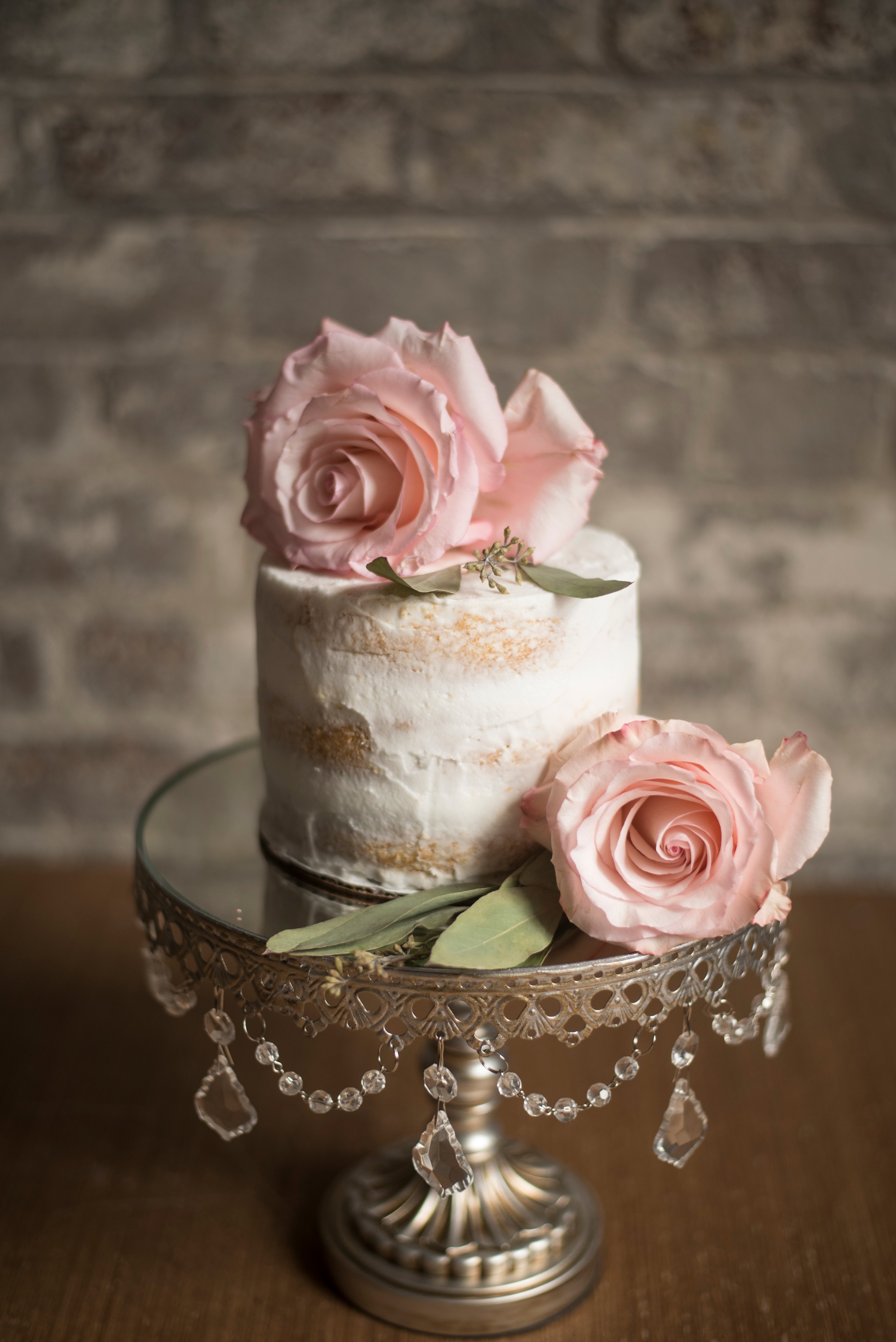Small white cake with pink roses on a decorative stand against a brick wall.
