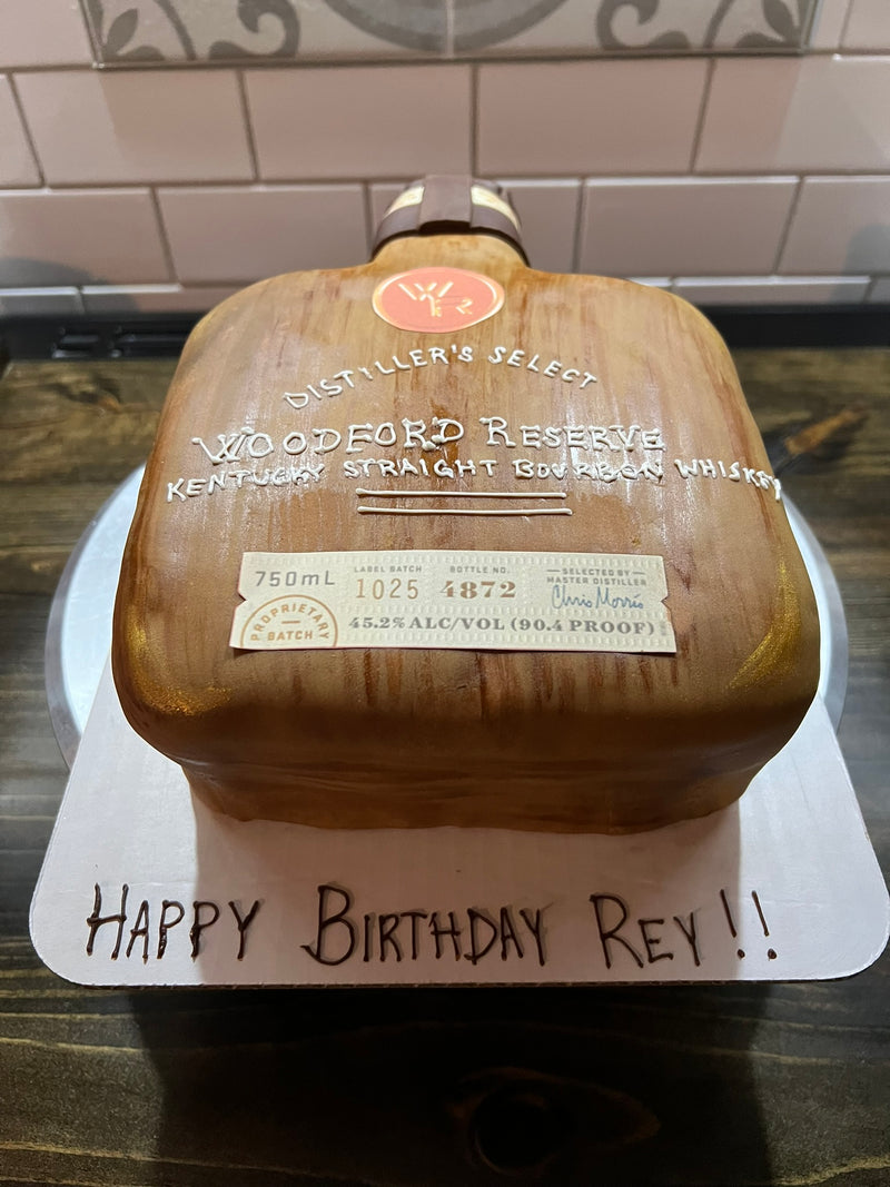 Wooden whiskey bottle-shaped cake with 'Woodford Reserve' branding on a white cake board.