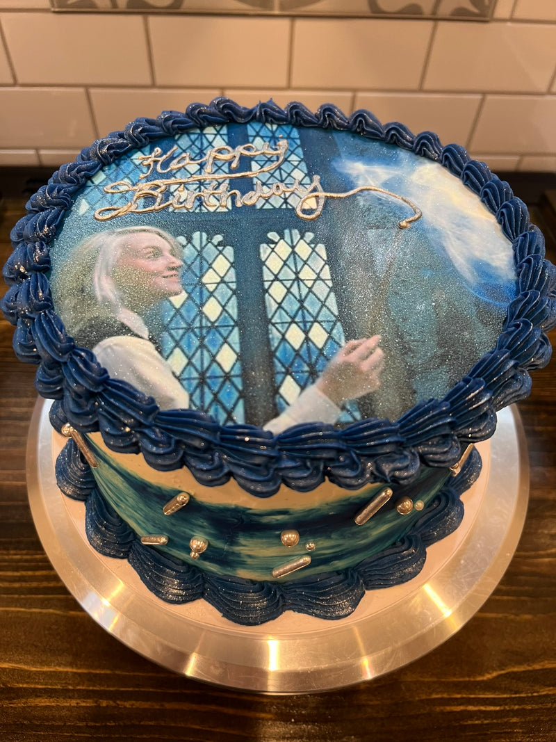 Decorative birthday cake with a scene of a man and woman by a stained glass window, on a wooden surface.
