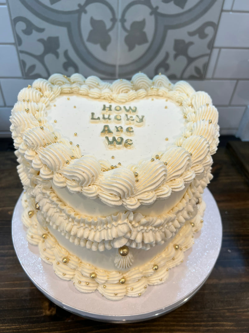 Two-tiered heart-shaped cake with 'How Lucky Are We' text on a decorative background.