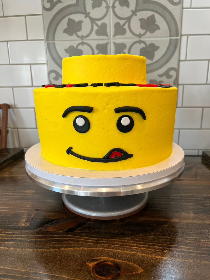 Yellow cake shaped like a Lego figure's head on a wooden table with a tiled wall background.