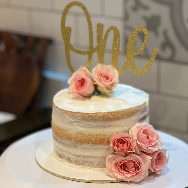 Small cake with pink roses and a gold 'One' topper on a white surface.