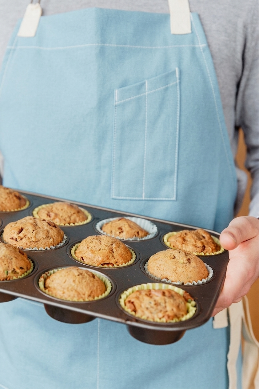 A person wearing a light blue apron holds a dark metal 12-cup muffin tray filled with baked muffins. The apron has a front pocket and white straps, and the person is wearing a gray long-sleeve shirt.
